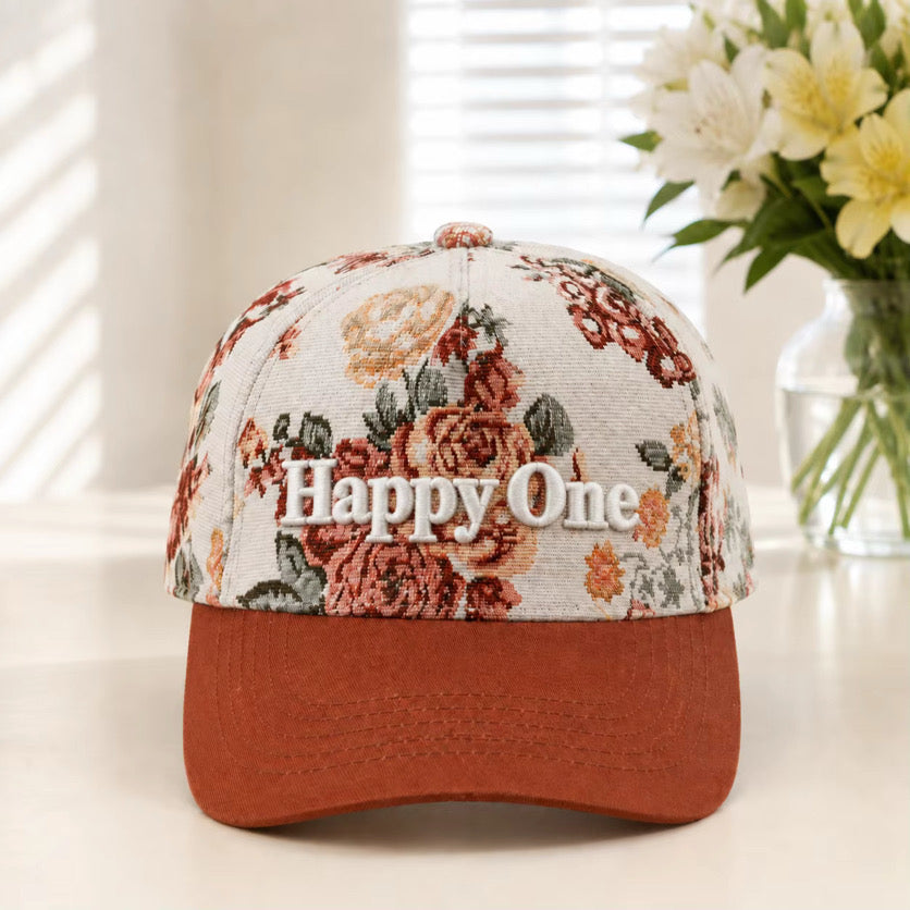 A baseball cap with a cream-colored floral patterned crown and a rust-orange curved brim. The front of the cap features the embroidered text 'Happy One' in white.