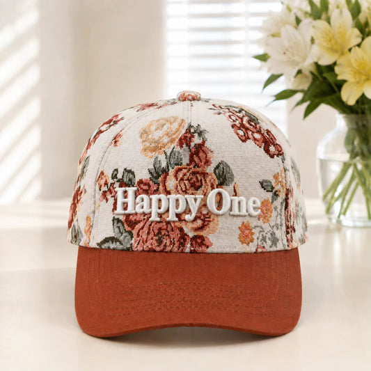 A baseball cap with a cream-colored floral patterned crown and a rust-orange curved brim. The front of the cap features the embroidered text 'Happy One' in white.
