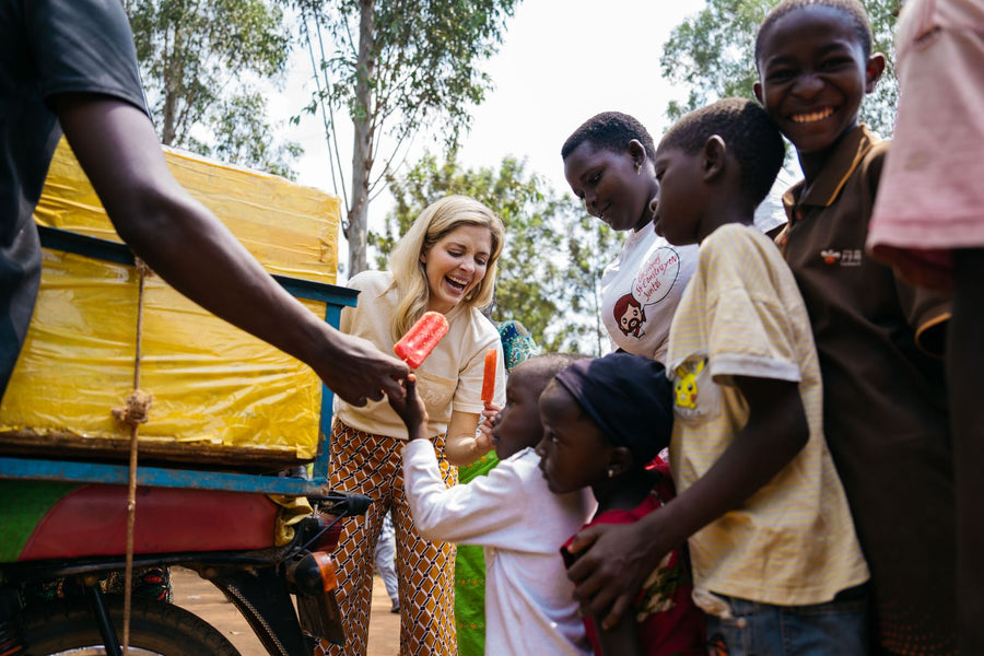 A woman smiling and handing a red popsicle to children gathered beside a yellow cart outdoors.